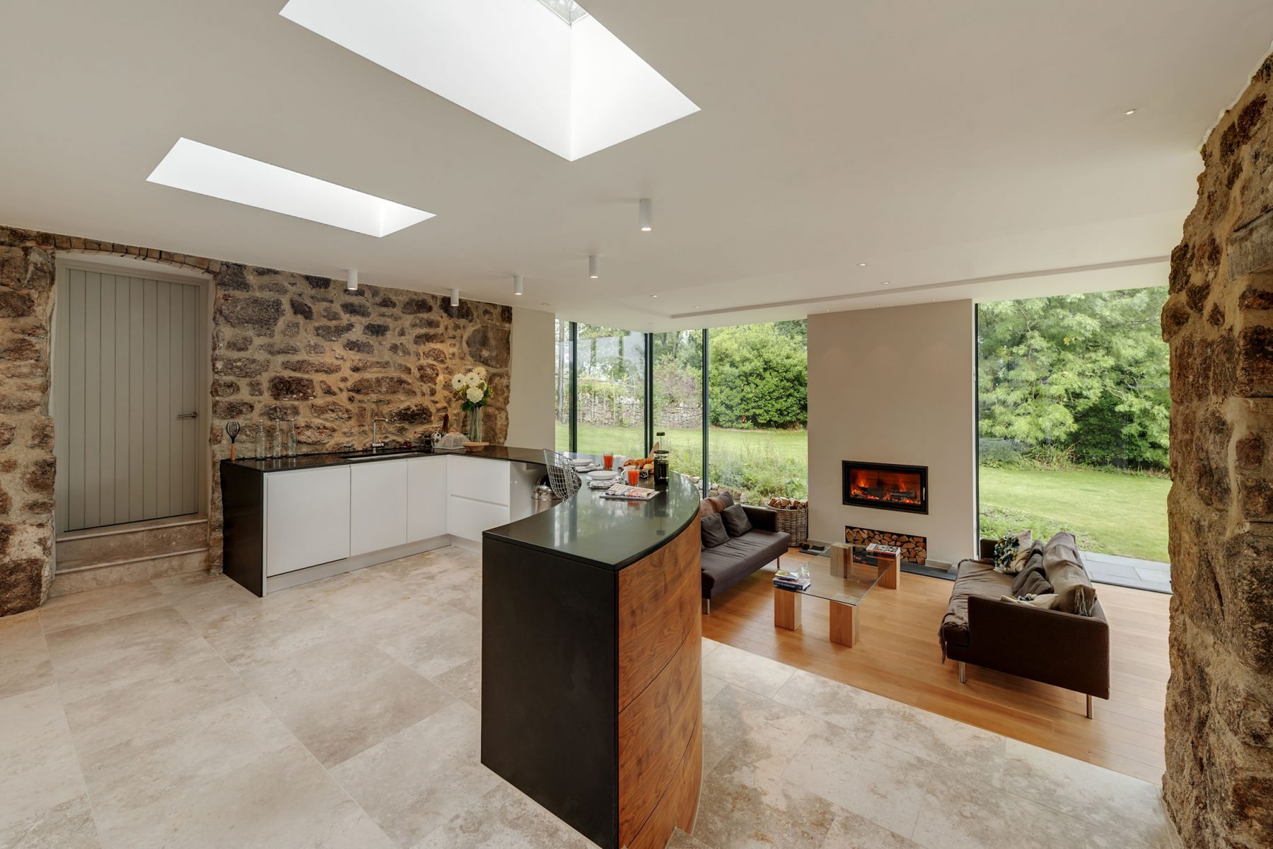 Curved kitchen island with generous flat roof lights above. Limestone floor, white kitchen cupboards with black worktop. Glass box living room beyond with inset wood burning stove.