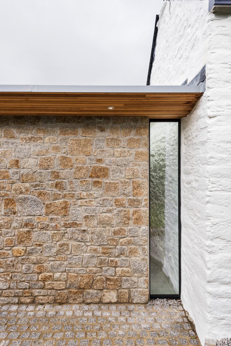 Granite stone wall with slot window and white painted stone of traditional Cornish cottage. Granite cobble setts for garden paving and western red cedar soffit to roof overhang.
