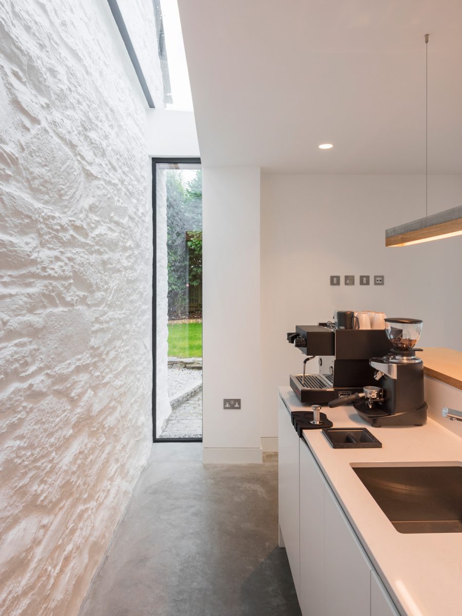 Coffee island joinery with concrete and timber strip pendant light. Vertical glass slot window with overhead slot roof light at abutment with cottage stone wall. Polished concrete floor and white painted stonework.
